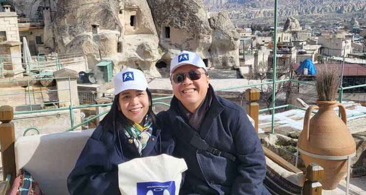 Pareja sonriente con gorras blancas sentada en un sofá de terraza al aire libre con las formaciones rocosas de Capadocia y los tejados del pueblo detrás de ellos