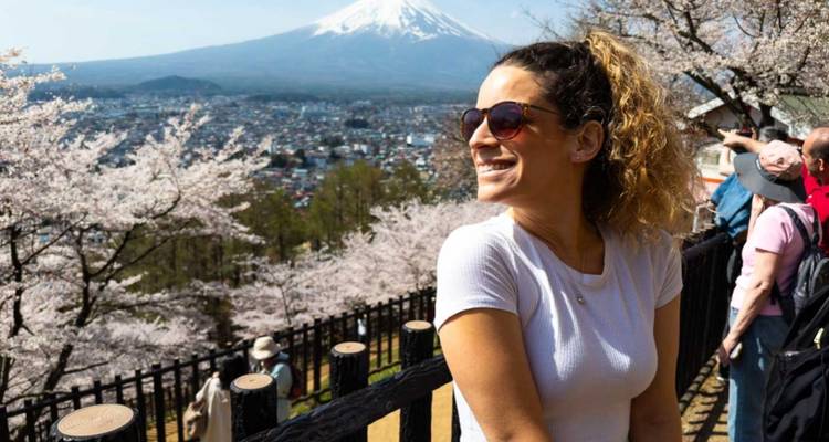 Smiling traveller surrounded by cherry blossoms with Mount Fuji towering behind and city below.