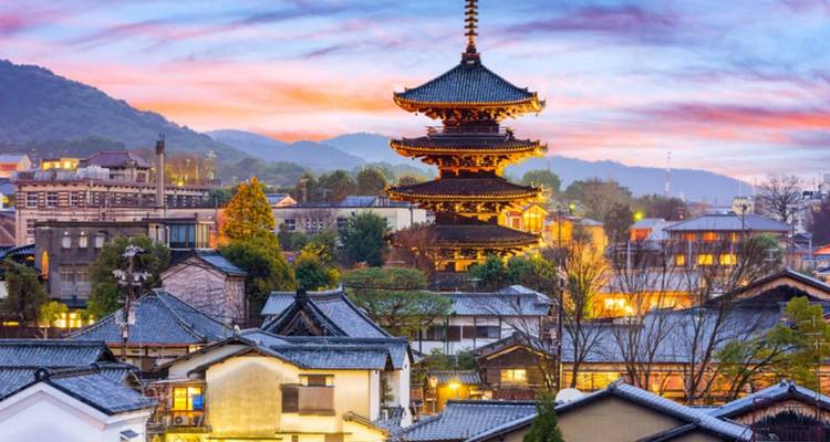 Scenic sunset view over traditional wooden pagoda rising above tiled rooftops with misty mountains in the background.