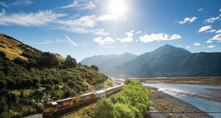 Ein Personenzug schlängelt sich durch ein weites Tal neben einem verzweigten Fluss mit sonnenbeschienenen Bergen im Hintergrund.