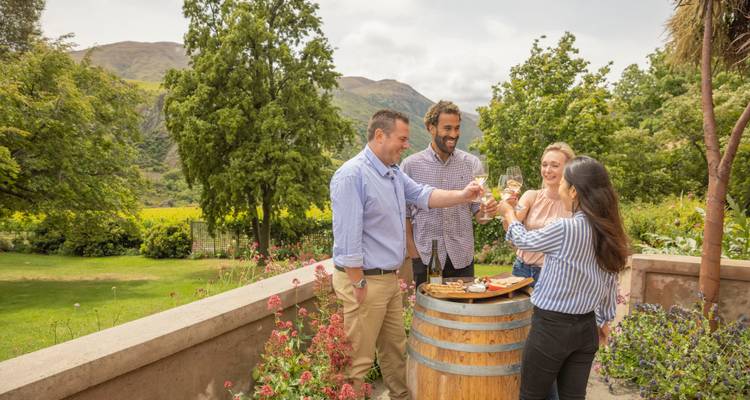 Gruppe von Freunden stößt mit Gläsern Weißwein neben einem Fass in einem üppigen Weinberggarten an.