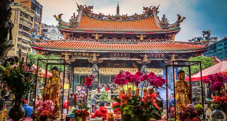 Cour de temple ornée remplie de fidèles, statues dorées et orchidées vibrantes sous un toit sculpté de manière complexe