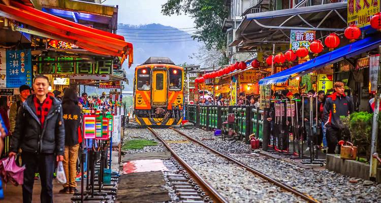 Train jaune roulant dans une rue étroite bordée de boutiques colorées éclairées par des lanternes et de visiteurs qui se promènent