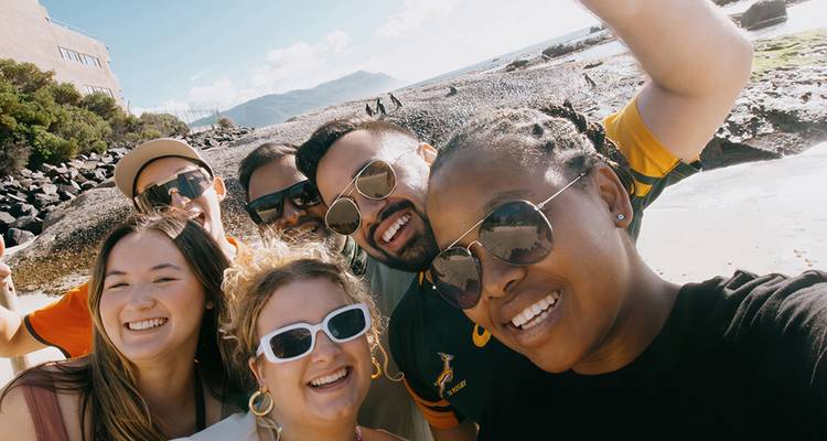 Una selfie grupal alegre en una costa rocosa con montañas de fondo.