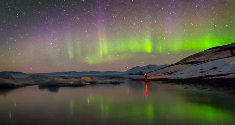 Aurora colorida reflejada sobre una laguna helada y tranquila bajo un cielo nocturno lleno de estrellas.