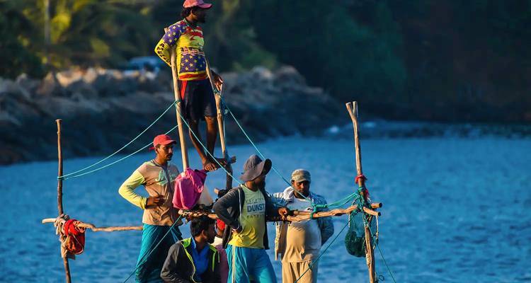 Pescadores en zancos equilibrándose en postes de madera sobre aguas costeras azul profundo durante la luz clara del día.