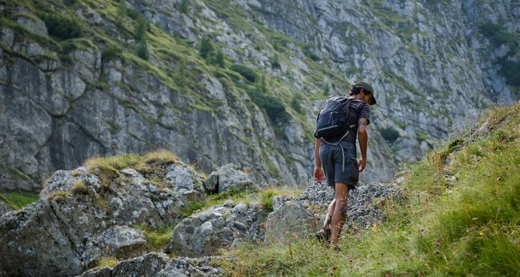 Rugzaktoerist die een rotsachtig alpenpad beklimt naast steile grijze rotswanden en groene hellingen.