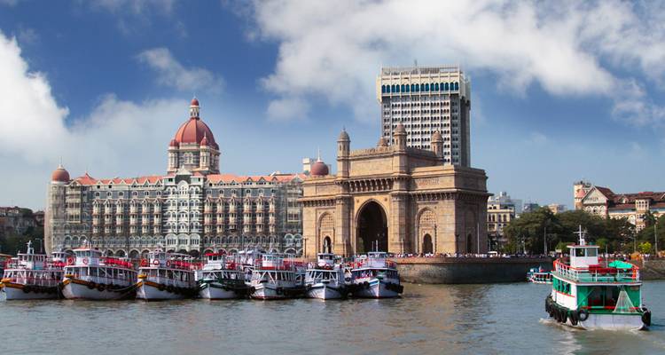 Los barcos están alineados en el puerto frente al arco de la Puerta de India y el Hotel Palacio Taj Mahal con cúpula bajo un cielo azul brillante.