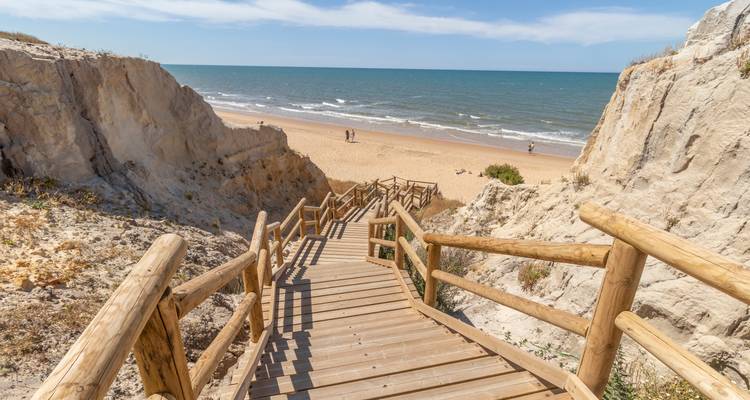 Houten boardwalk die naar beneden leidt naar een rustig strand met zachte golven onder een zonnige hemel.