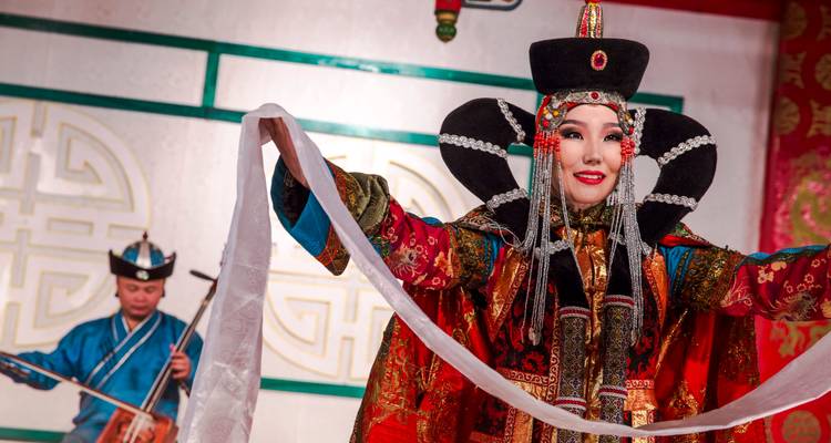 Vibrantly dressed Mongolian dancer greets the audience holding a white ceremonial scarf as a musician plays behind.