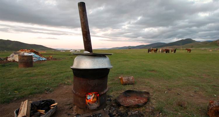 Rustic metal stove with open fire in a wide grassy steppe at dusk, horses in the distance under cloudy sky.