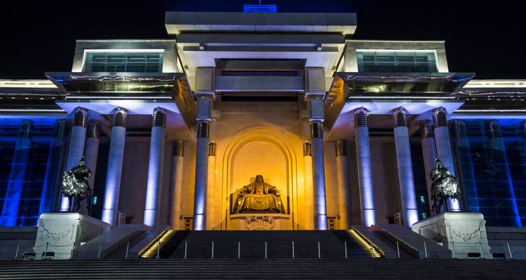Illuminated Government Palace and seated Genghis Khan statue in Ulaanbaatar glowing blue and gold at night.