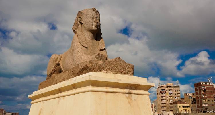 Standbeeld van een sfinx met een dramatische bewolkte hemel en stedelijke gebouwen op de achtergrond.