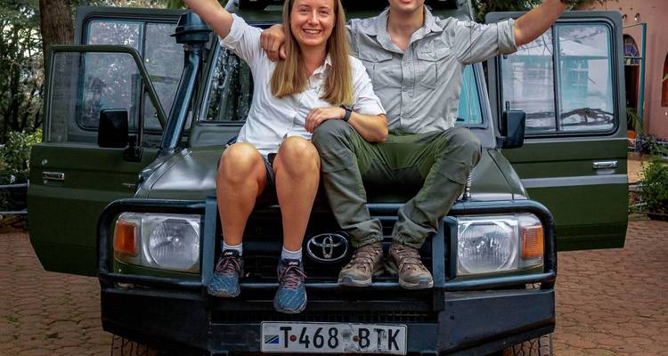 Zwei Personen sitzen auf der Motorhaube eines Safarifahrzeugs und lächeln.