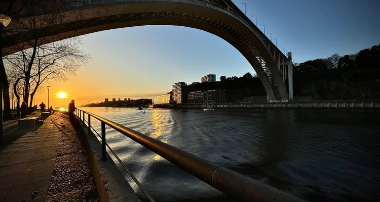 Sunset view under the Arrábida Bridge in Porto with the skyline in the background.