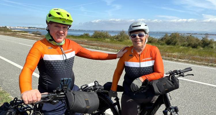 Two cyclists on a road by a scenic landscape with water and greenery.