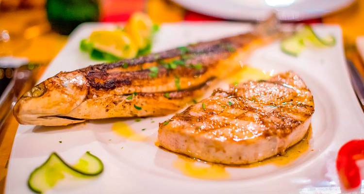 Close-up van een gegrilde hele vis en een tonijnsteak bedruppeld met olijfolie op een restaurantbord.