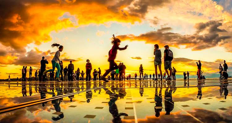 Silhouetten van mensen weerspiegeld op een glazen zonne-installatie bij zonsondergang met dramatische oranje wolken.
