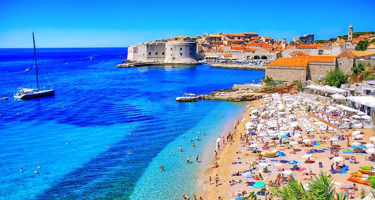 Druk kiezelstrand met parasols en zwemmers met uitzicht op Dubrovniks middeleeuwse muren en helder turquoise water.
