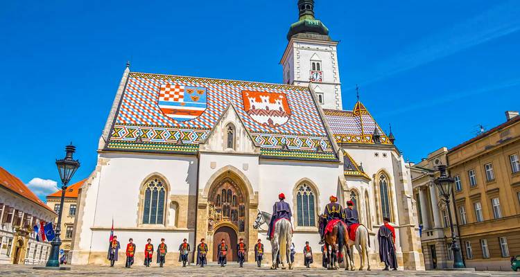 Ceremonial guards on horseback in front of Zagreb’s multicoloured-tiled St. Mark’s Church under blue skies.