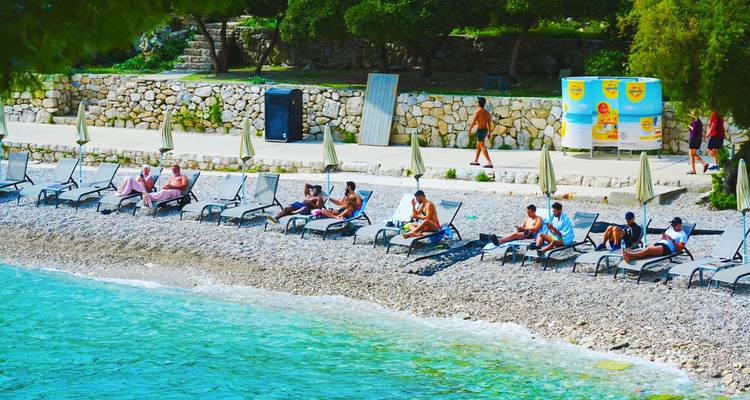 Relaxed sunbathers on loungers along a pebbly Adriatic beach with clear turquoise water.