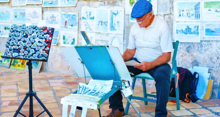 Elderly artist sketching seaside scenes at an outdoor stall decorated with watercolour paintings.