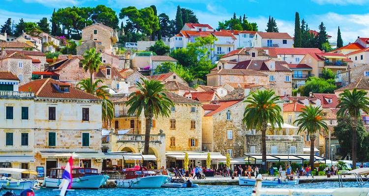 Charming harbour lined with stone houses, palms and cafés; boats moored in bright blue water.
