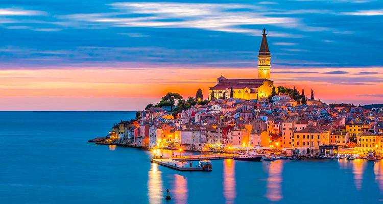 Sunset over the colourful hilltop town of Rovinj with its landmark church tower reflected in calm water.