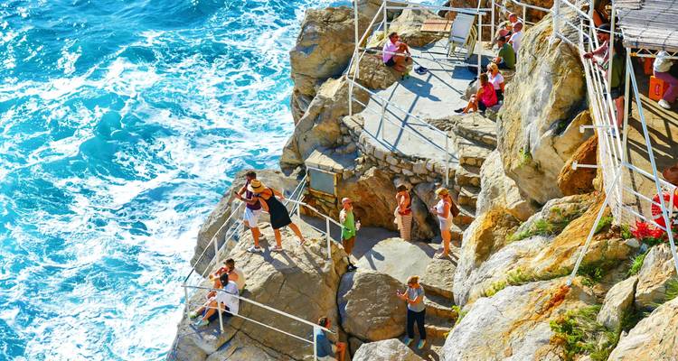 Visitors perch on rocky terraces of a seaside bar overlooking crashing turquoise waves.