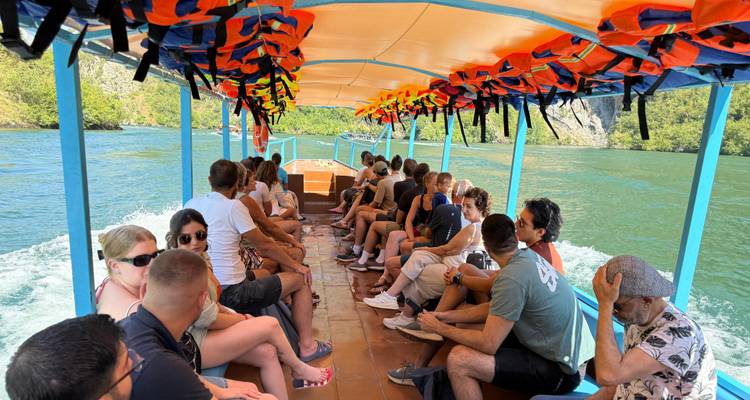 Toeristen gezeten op een open boot die vaart langs een groen meer met reddingsvesten boven hun hoofd.
