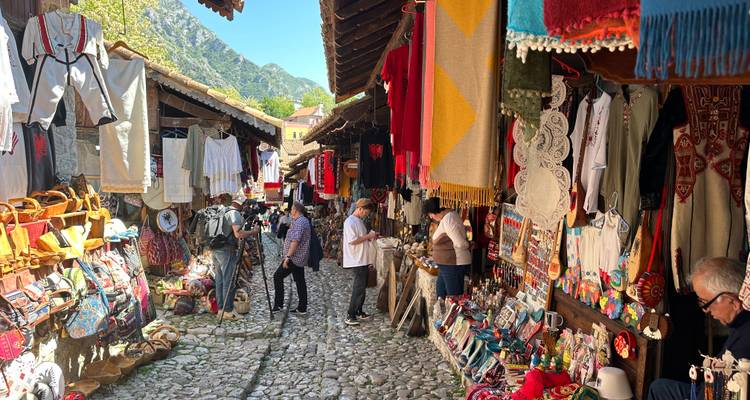 Keienstenenbazaar omzoomd met kleurrijke textiel, ambachtelijke producten en winkelende mensen in een historische Albanese stad.