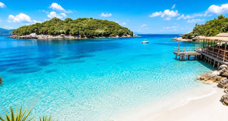 Playa de arena blanca impecable con agua turquesa cristalina y un islote exuberante y verde en Ksamil.