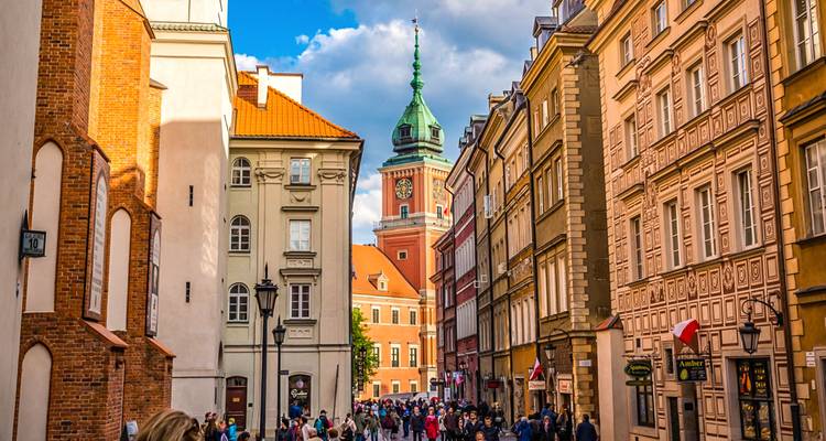 Bustling pedestrian street lined with colorful buildings leading to a tall clock tower in Warsaw's Old Town.