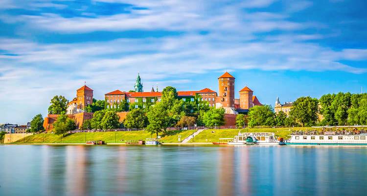 Wawel Castle perched above the Vistula River with reflections shimmering in calm water under a bright sky.