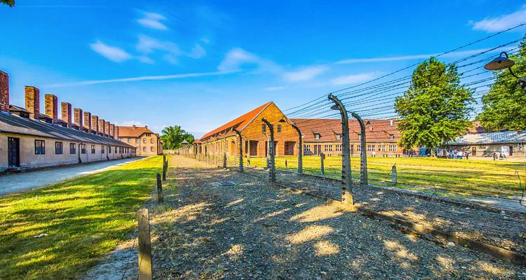 Barbed wire fences and brick barracks of the Auschwitz concentration camp under a clear blue sky.