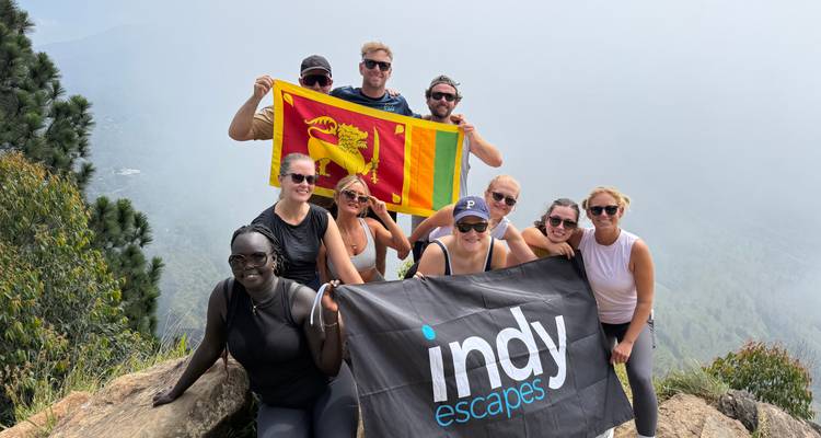 Un grupo de senderismo celebra en la cima brumosa sosteniendo la bandera de Sri Lanka y un banner del tour.