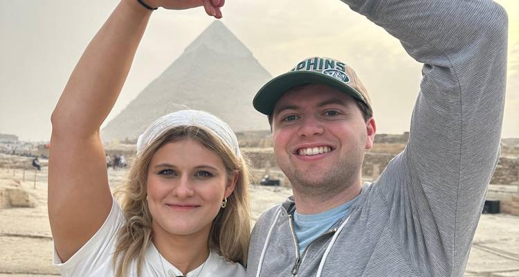 Smiling couple posing with arms forming a pyramid shape in front of the Great Pyramid in hazy light.