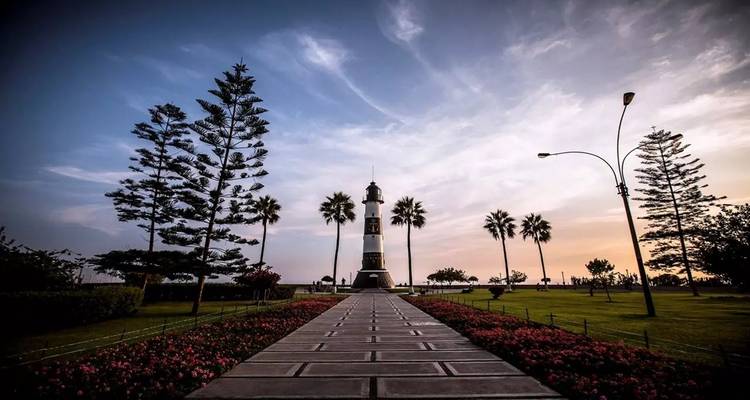 Pathway lined with flowers leading to a lighthouse and tall palm trees at dusk in Lima.