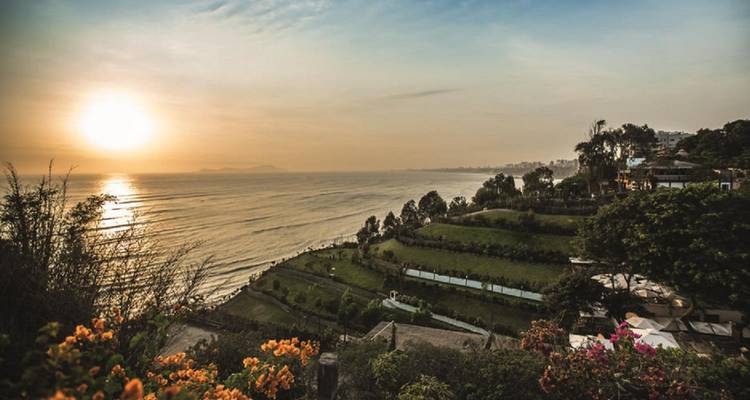 Sun setting over the Pacific coastline of Lima, casting warm light on terraced parks and the ocean.