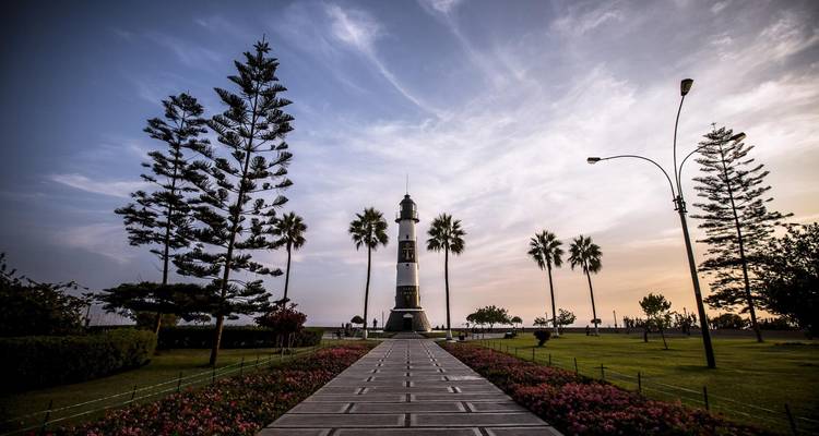 Path lined with flowers leading to the Faro la Marina lighthouse framed by palm trees at dusk.