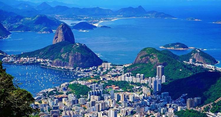 Hoher Panoramablick auf Rio de Janeiro mit dem Zuckerhut, grün getupften Gipfeln, der tiefblauen Guanabara-Bucht und der dicht gedrängten Stadtsilhouette unter einem klaren Himmel.