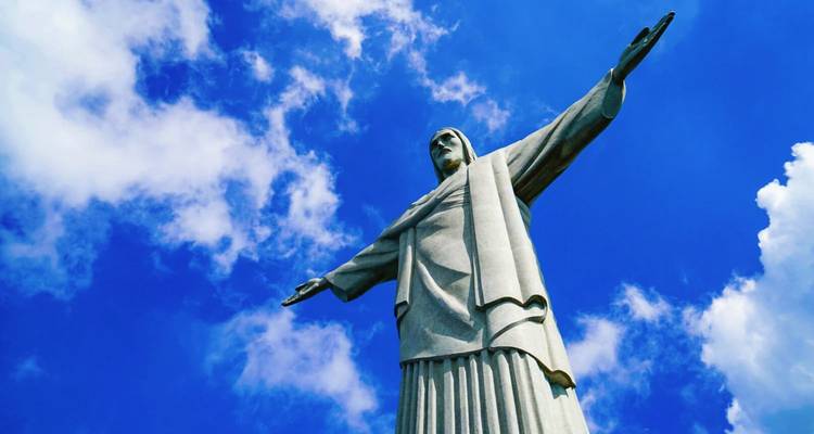 Die ikonische Christus-Erlöser-Statue steht hoch aufragend vor einem strahlend blauen Himmel mit vereinzelten Wolken, die Arme ausgestreckt.