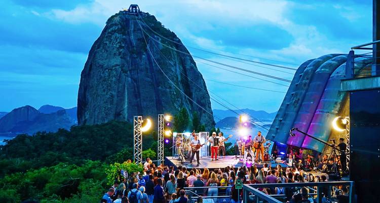 Ein lebhaftes Freiluftkonzert findet auf einer Plattform in der Nähe der Seilbahnstation am Zuckerhut statt, bunte Bühnenlichter leuchten, während der Abend hereinbricht.