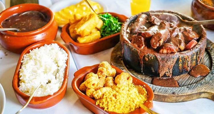 Traditionelle brasilianische Gerichte wie Feijoada, Reis, Bohnen und Farofa werden in rustikalen Tontöpfen auf einem weiß gedeckten Tisch serviert.