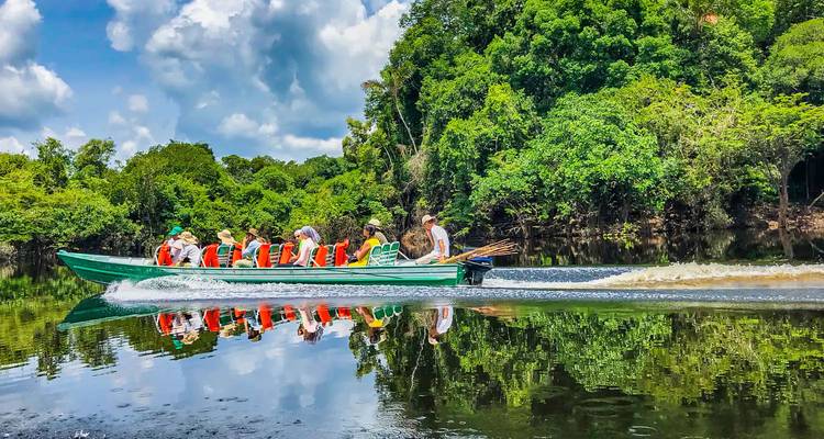 Una lancha motora verde y rápida que lleva turistas con chalecos salvavidas avanza velozmente por un río selvático de aguas tranquilas como un espejo, con el exuberante bosque reflejado en el agua.