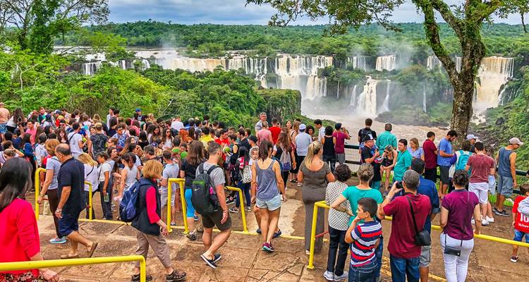Große Menschenmengen säumen die Geländer mit Blick auf die dramatischen Vorhänge der Iguazu-Wasserfälle, umgeben von üppigem Grün.