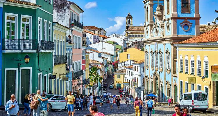 Eine lebhafte Kolonialstraße mit bonbonfarbenen Fassaden, Balkonen und Kirchtürmen summt vor Fußgängern unter einem strahlend blauen Himmel.