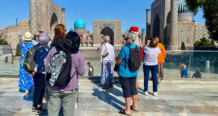 A tour group observes the grand turquoise domes and mosaic facades of Registan Square under a clear sky.