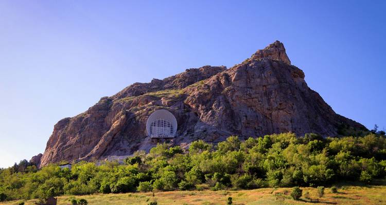 A rugged rocky mountain with a distinctive cave-like monument set above green foothills and blue sky.