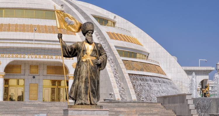 A golden statue of a Turkmen leader holding a flag stands before a grand marble museum with cascading fountains.